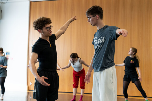 Two young students wearing glasses and dance clothing are working together. They have their arms outstretched to the side and are speaking to each other. In the background there are three other dancers