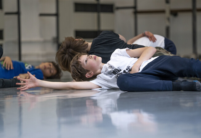 Children in a dance studio wearing exercise clothes lying on a dance studio stretching their arms out