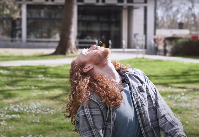 A person with long ginger hair sitting on some grass. They are leaning their head back and blowing a Malteaser in the air with their mouth.