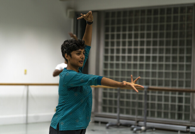 Bharatanatyam teacher, Shivaangee Agrawal, wears a turquoise top and has short black hair. Their right arm is completely outstretched in front of their body, palm facing up and fingers splayed. Their left are is above their head, two fingers and thumb pressed together in a classical mudra position.