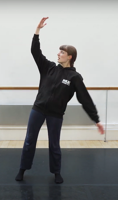 Person in a dance studio wearing a black hoodie and black trousers. They are bending one arm over their head and have their other arm down by their side.