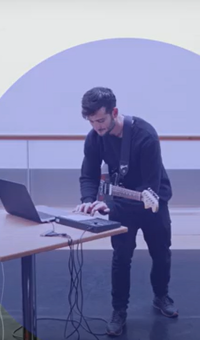 Person with a guitar in a dance studio taps into a keyboard. There is a blue opaque circle round them.