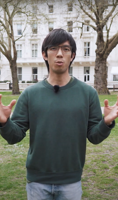 Person in an outside environment wearing a green jumper and jeans. They are holding their hands in front of them.