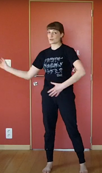 Person in a room with a red wall and wooden floor. They are wearing a black t-shirt and black trousers and are moving their arms in front of them.