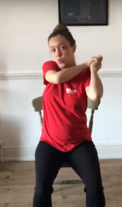 Person in a red t-shirt and black trousers sitting on a chair in a living room. Their hands are held together and they are swinging them over their right shoulder.