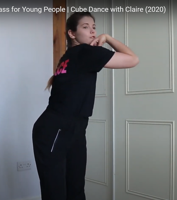 Person in a white room wearing a black t-shirt and black trousers. They are pointing their elbow to their left shoulder.