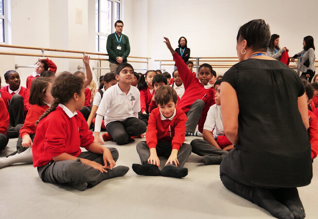 Primary school pupils sitting on the floor of dance floor listening to a teacher