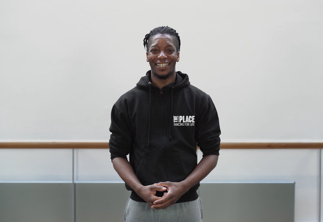 A person standing in a dance studio with a ballet barre behind them. They are wearing a black hoody with The Place logo on in the top right corner