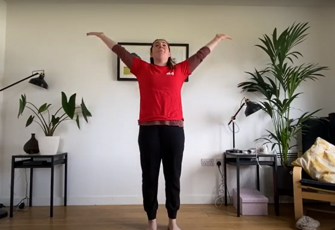 Dance teacher in their lounge wearing a red t-shirt and black trousers stretching their arms upwards towards the ceiling.