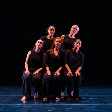 5 women all dressed black, 3 are seated with two standing behind them, all leaning to the right