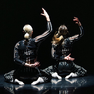 Two figures wearing striped body suits, face away from the camera, with one arm raised, the other behind their backs