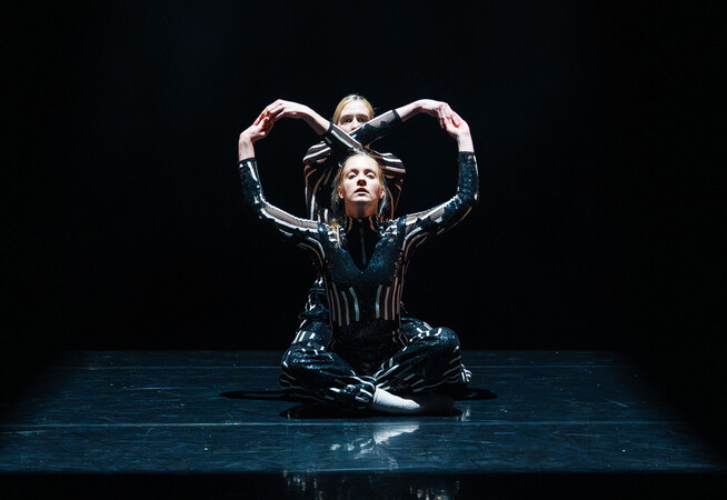 Two figures in striped body suits on a black background, sit on the floor facing forwards. Their arms make two circles