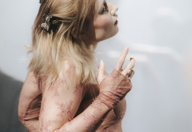 A person with long blond hair and lacey top stands sideways to the camera, looking slightly upwards