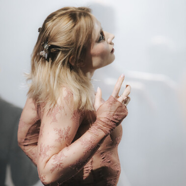 A person with long blond hair and lacey top stands sideways to the camera, looking slightly upwards