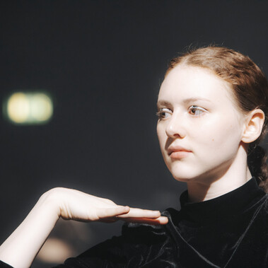 A person with dark ginger hair, wearing a black high neck top stares past the camera.