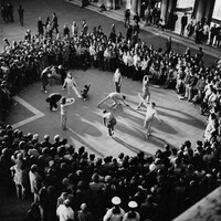 Black and white image of Merce Cunningham's Event performed in Venice's St Mark's Square in 1972. The image is taken from a bird's eye view. 7 dancers wearing light colour costumes and 5 wearing darker costumes and encircled by a large audience.