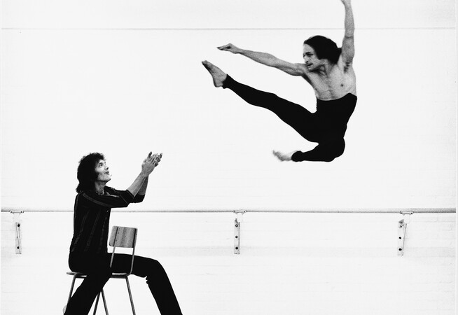 Black and White photo of Robert (Bob) Cohan in rehearsal with a member of the London Contemporary Dance Theatre. Cohan is on a chair clapping at a performer in the air with black leggings and kicking his leg forward