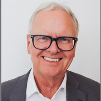 Portrait image of Alan Bishop, a white man seen smiling wearing dark square glasses, a shirt and a blazer. Alan has white hair