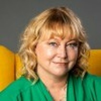 A blond woman with mid-length hair is looking in the camera and smiling. She is wearing a green top and is sat on a yellow chair.