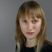 A white blond person is looking in the camera in front of a grey background. They have long blond hair and a fringe and are wearing a black t-shirt.