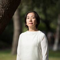 a lady is standing in a garden wearing a white top, looking just above at the camera. She has short brown hair.