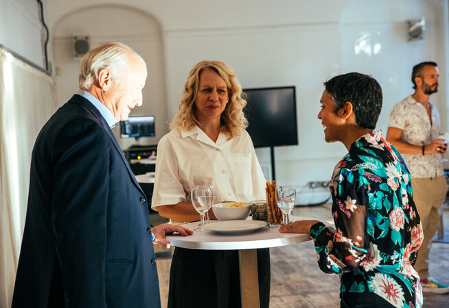 Patron Lord Simon Russel of Liverpool, Chief Executive Clare Connor and Governor Eva Sarma are chatting  standing up around a tall table.