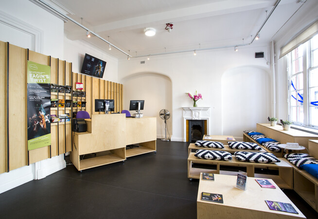 fish eye view of The Place foyer and Box Office area with wooden benches with cushions on them facing wooden tables and a modular wooden box office desks with computers. The background of the box office area is wooden and is displaying posters.