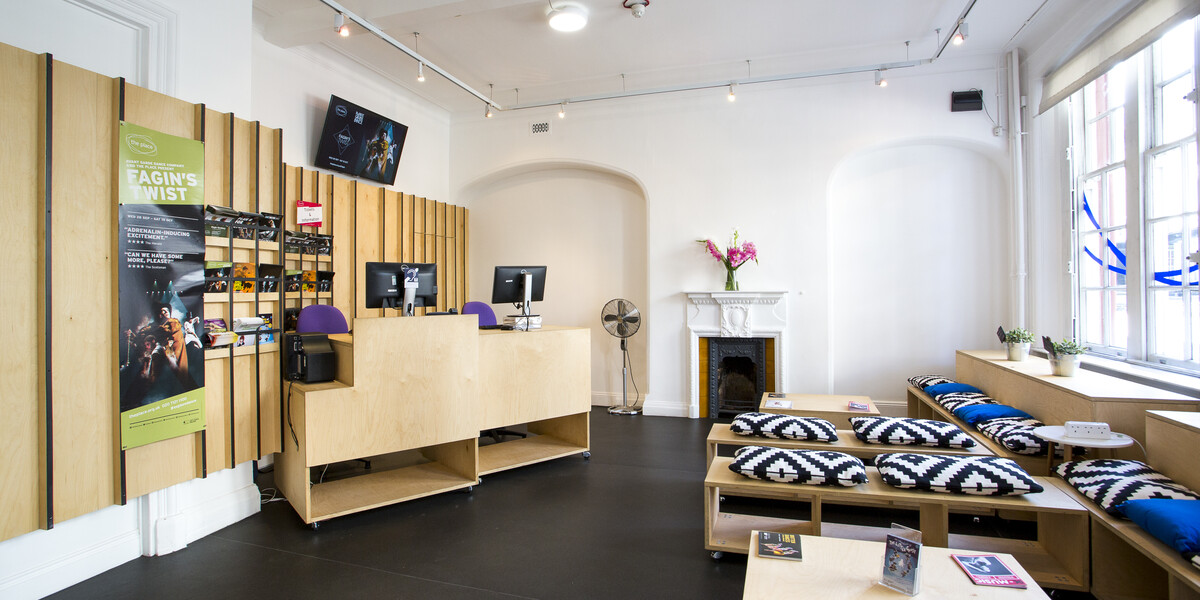 fish eye view of The Place foyer and Box Office area with wooden benches with cushions on them facing wooden tables and a modular wooden box office desks with computers. The background of the box office area is wooden and is displaying posters.