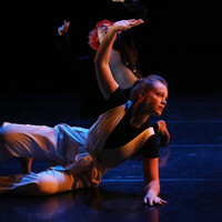 Image of Isis Clunie on a stage. Stage is darkly lit. Isis is wearing a blue shirt with white trousers. Isis lies on the floor with their left arm supporting them. Their right hand is raised up above them.