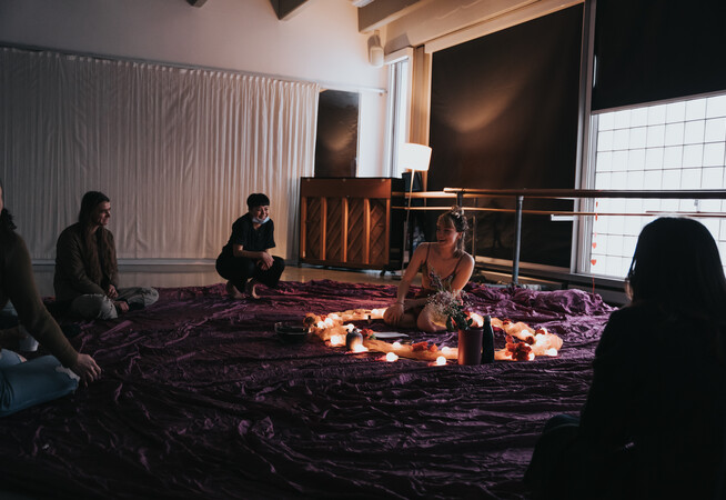 On some deep purple fabric is a circle made up of light fabric, fairy lights and roses. A person wearing a mesh cropped top and purple silk shorts kneels, their arms in out in front of them, elbows slightly bent and palms facing the body. Between their knees is a sheet of paper and in front of this is a bowl. To the left is a pink vase of flowers.