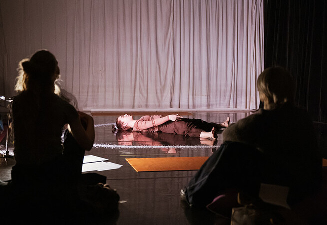 A person in the background lays on the black floor with a white ring of salt around their body and a spotlight on them, they are wearing burgundy clothes and their are two people watching them in the foreground