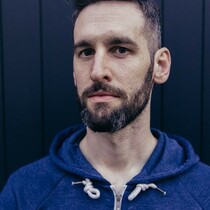 A man with short black hair and a beard is wearing a blue zip up hoodies and is looking into the camera with a serious expression