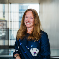 A woman with light red hair and a big smile looks at the camera. She is leaning on the metal railing and wearing a blue silk long sleeved top