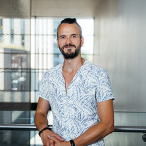 A man with short black hair and a black beard looks at the camera with a slight smile. He is wearing a light blue and white patterned shirt and is leaning on a railing with his hand clasped together