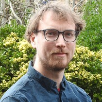 A man wearing a dark blue shirt and blue framed glasses looks into the camera. In the background there are green bushes and trees, he has light brown hair and a beard