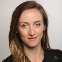 A woman with long light brown hair, wearing a black short sleeved top smiles and looks into the camera, there is a white background behind her