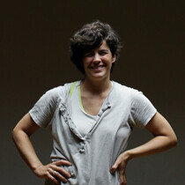 A woman wearing a baggy white t-shirt with short dark hair smiles at the camera with her hands on her hips. There is a dark background behind her