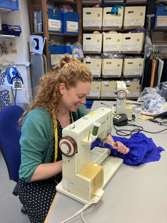 A woman leans over a sewing maching, she has a tape measure around her neck and she is smiling