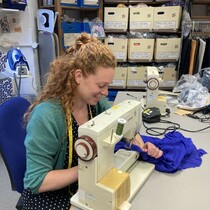 A woman leans over a sewing maching, she has a tape measure around her neck and she is smiling