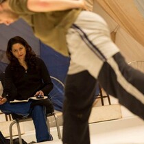 A woman sits on a chair with a notebook, she is watching a dancer in during a rehearsal, she has an intense look on her face