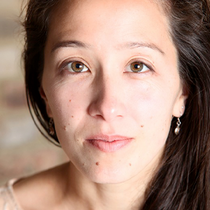 A woman with brown eyes looks directly into the camera, she is wearing a cream top and had brown long hair