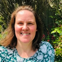 A woman with mid length brown hair is smiling and looking at the camera. She is wearing a green and white floral top and behind her there are plants and flowers.