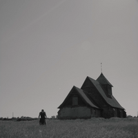A black and white photo with a large house on the right hand side and a woman can be seen on the left in the long grass, she is looking down