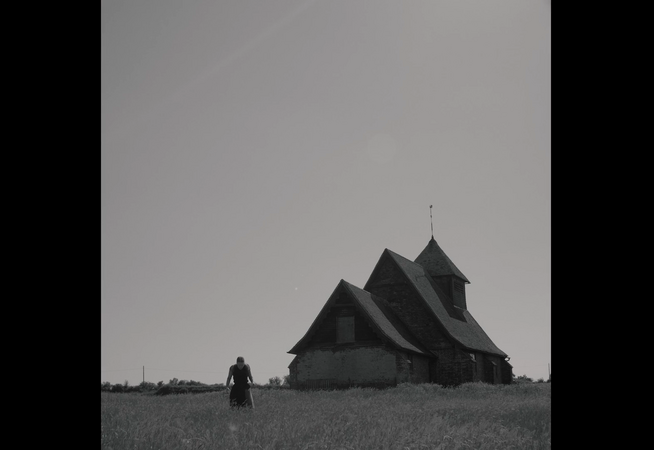 A black and white photo with a large house on the right hand side and a woman can be seen on the left in the long grass, she is looking down