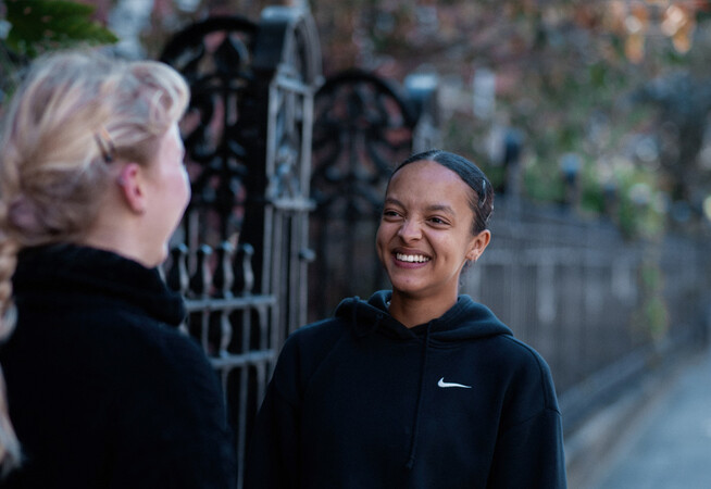 Two students stand outside LCDS, there is a blurred background that has a metal fence and trees in, one students is smiling at the other