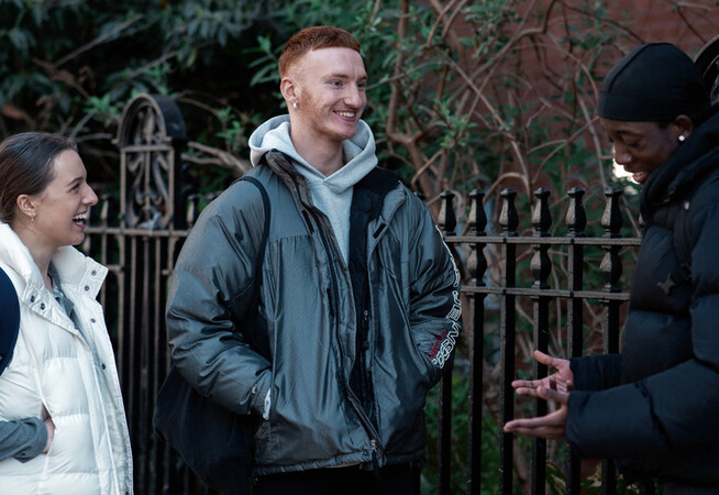Three LCDS students stand outside The Place, there is a metal fence and trees behind them, one student is making the other two laugh