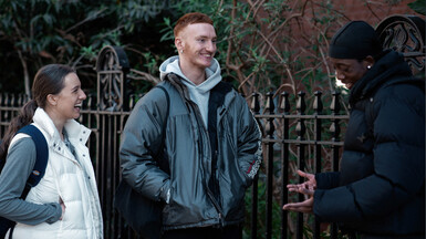 Three LCDS students stand outside The Place, there is a metal fence and trees behind them, one student is making the other two laugh