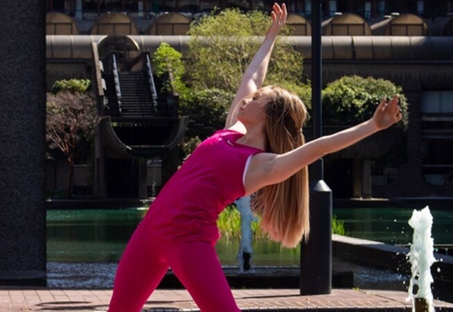 Image of Ellie dancing centrally in the camera. Behind Ellie is a water feature and a small tree. Ellie is wearing all pink clothes and has long blonde hair.