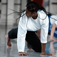 A person wearing a white long sleeved top in our studio. They are on the floor with their arms are stretched to the floor and their head is looking to the right, which makes their hair braids fly out