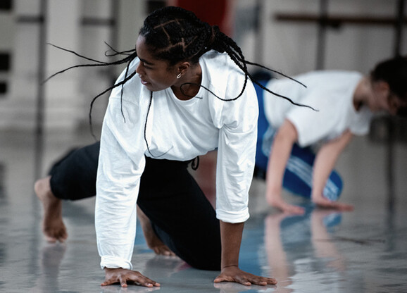 A person wearing a white long sleeved top in our studio. They are on the floor with their arms are stretched to the floor and their head is looking to the right, which makes their hair braids fly out